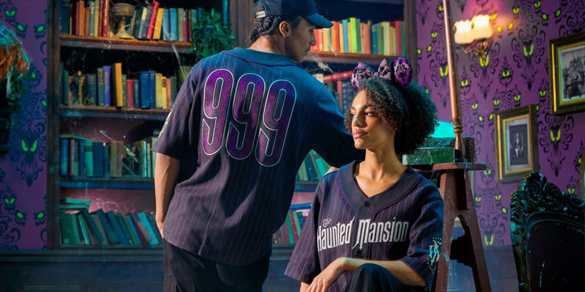 Two people in a spooky, purple-lit library sport Haunted Mansion 2025 merch jerseys—one with "999" on the back, the other with "Haunted Mansion" on the front. The woman also wears sparkly cat ears. Green eyes decorate the wallpaper.