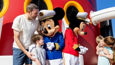 A family with two children meets Mickey and Minnie Mouse in sailor outfits on a cruise ship deck, with bright red and yellow colors in the background.