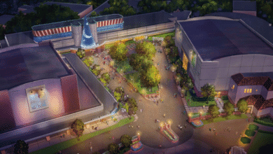 Aerial view of a theme park plaza at dusk, featuring a central open area with trees, pathways, people walking, and large buildings surrounding the plaza, including one with a blue sorcerer hat and the Star Wars Launch Bay nearby.