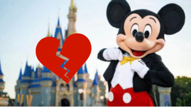 Mickey Mouse stands in front of a castle at a theme park, smiling and pointing at his bow tie. A large red broken heart graphic is superimposed over the image.