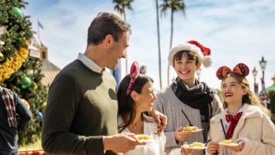 A group of four people, some wearing festive hats and mouse ears, smile and hold plates of food near a decorated Christmas tree outdoors on a sunny day. Palm trees are visible in the background.