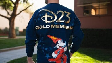 A person outdoors wears a navy blue jacket featuring “D23 Gold Member” in gold letters and an image of Mickey Mouse as the Sorcerer’s Apprentice from Fantasia on the back.