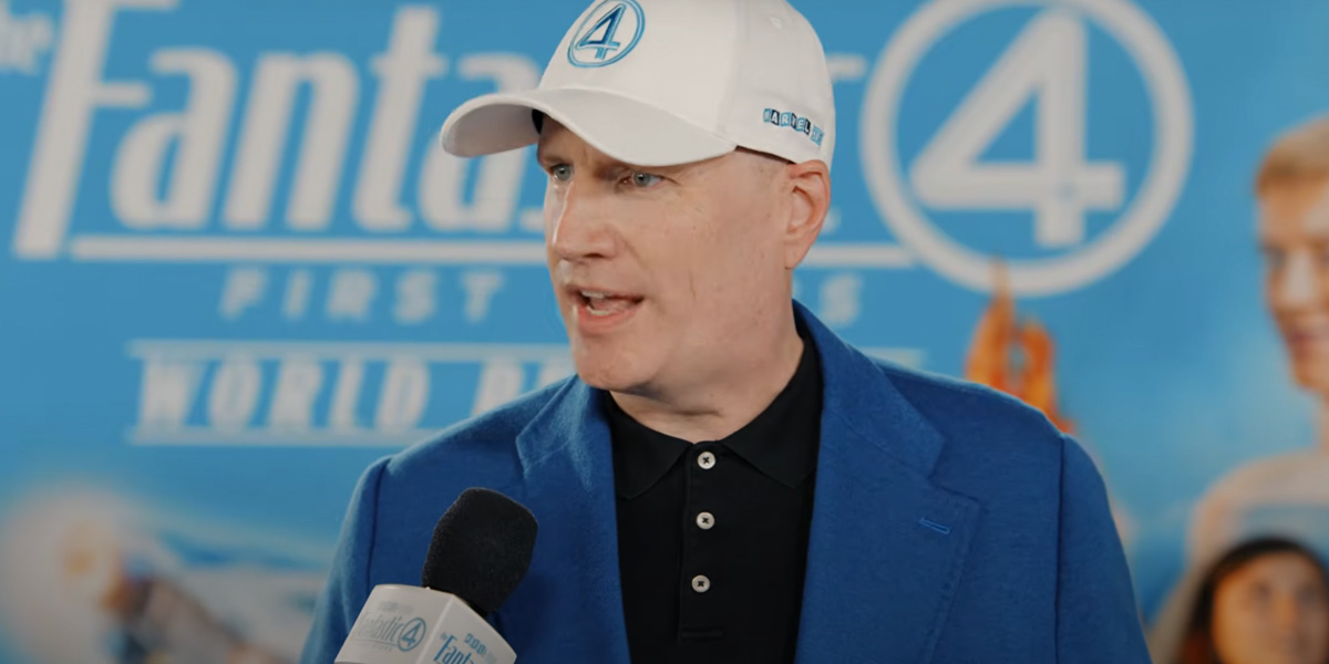 Kevin Feige in a white Fantastic 4 cap speaks into a microphone at a “Fantastic Four” event, with a blue promotional backdrop featuring the movie’s logo behind him.