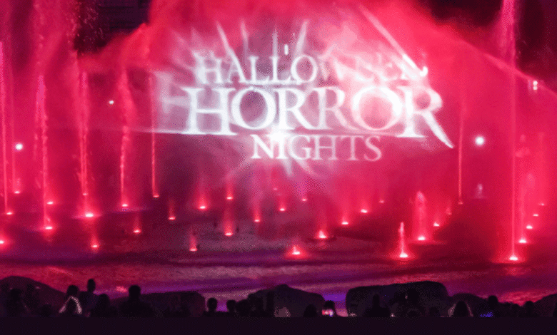 Red and purple fountains spray water in front of a projected Halloween Horror Nights logo, as a crowd of silhouetted people gathers to watch the spooky nighttime show at Halloween Horror Nights 34.
