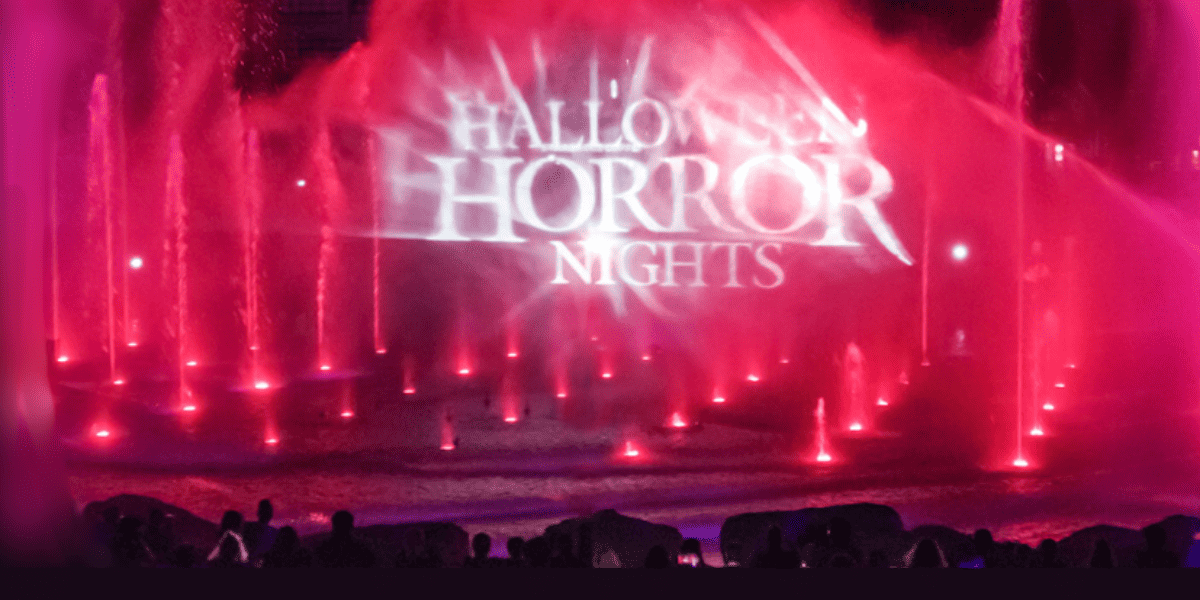 Red and purple fountains spray water in front of a projected Halloween Horror Nights logo, as a crowd of silhouetted people gathers to watch the spooky nighttime show.