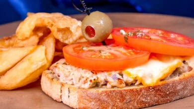 An open-faced tuna melt sandwich topped with melted cheese and fresh tomato slices, garnished with a green olive on a toothpick, served with thick-cut potato wedges on a wooden plate.