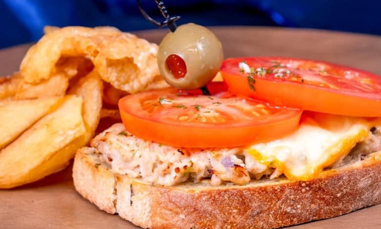 An open-faced tuna melt sandwich topped with melted cheese and fresh tomato slices, garnished with a green olive on a toothpick, served with thick-cut potato wedges on a wooden plate.