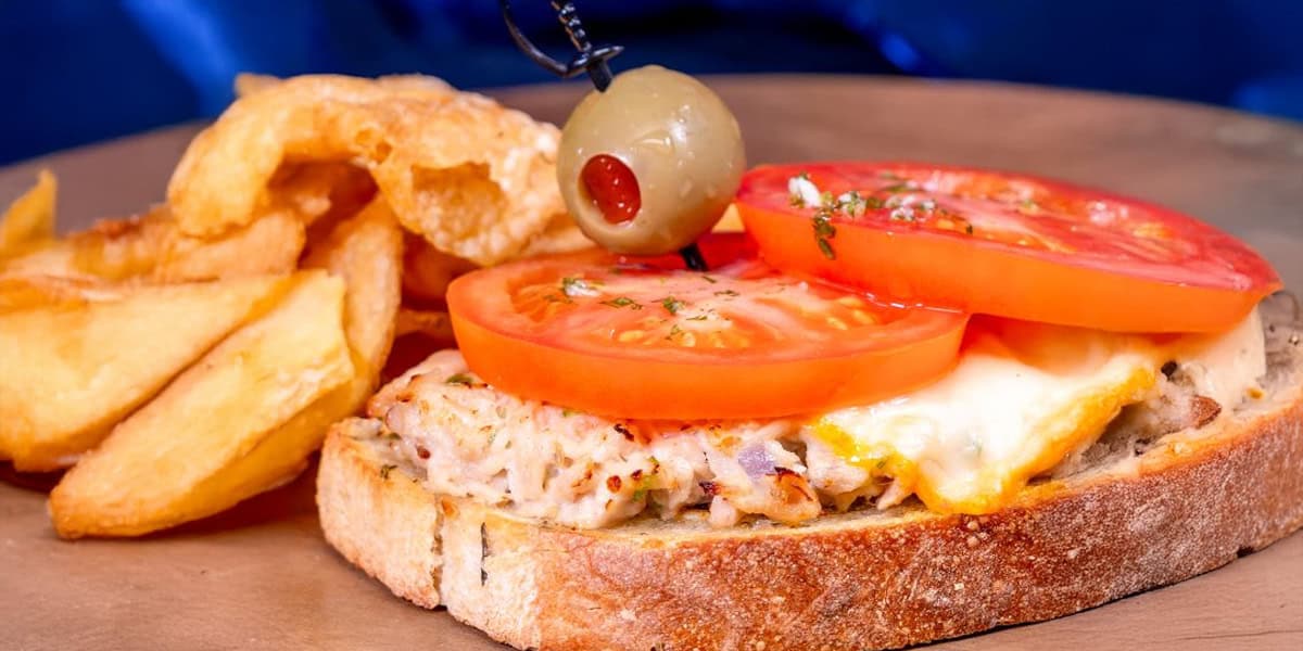 An open-faced tuna melt sandwich topped with melted cheese and fresh tomato slices, garnished with a green olive on a toothpick, served with thick-cut potato wedges on a wooden plate.