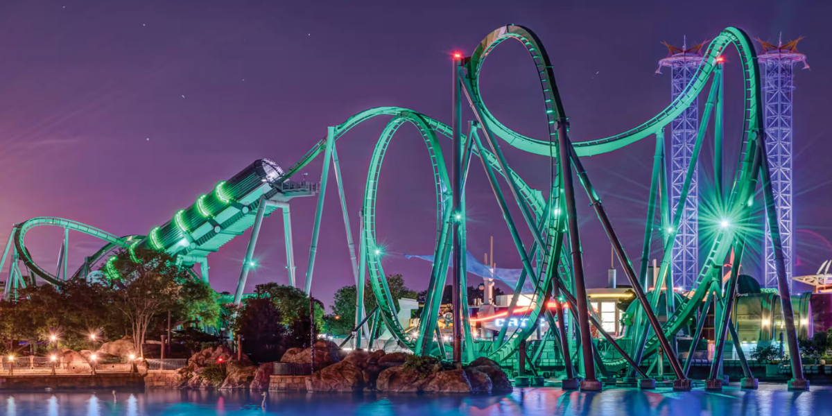 A brightly lit roller coaster with green tracks twists and loops against a purple night sky, with water and rocks in the foreground and amusement park buildings in the background.