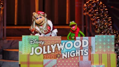 Miss Piggy and Kermit the Frog stand behind a stack of colorful presents with a sign reading “Disney Jollywood Nights 2025,” surrounded by holiday decorations and warm lights.