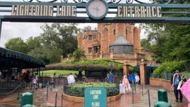 Entrance to the Haunted Mansion ride at a theme park, with a sign reading "Lightning Lane Entrance" and guests wearing rain ponchos walking past the ornate, spooky brick building in the background.