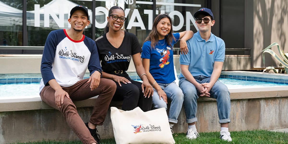 Four smiling people sit together outdoors by a shallow pool, wearing themed shirts and hats. A large canvas tote bag with a cartoon logo rests in front of them. The word "INNOVATION" is visible on the building behind them.