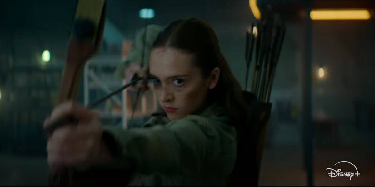 A young woman with brown hair aims a drawn bow and arrow intensely, appearing focused and determined. She has a quiver of arrows on her back. The scene is dimly lit, with a blurred background and a Disney+ logo in the corner.