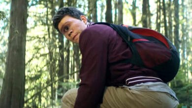 A young man wearing a maroon jacket and backpack crouches in a forest, looking back over his shoulder with a concerned expression. Sunlight filters through the tall trees in the background.