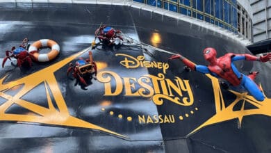 Statues of Spider-Man and two spider-robots cling to the side of a ship with “Disney Destiny Nassau” written in gold letters. One spider-robot holds a life preserver, and the background shows part of the ship's structure.