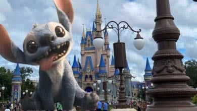 A large, animated Stitch character from Disney's "Lilo & Stitch" is superimposed in front of Cinderella Castle at Disney World, with visitors walking around under a partly cloudy sky.