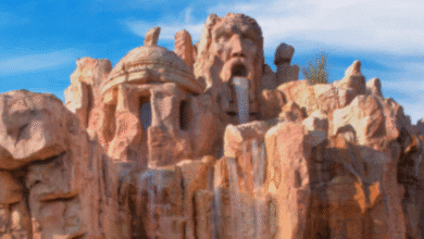 A large universal rock formation sculpted to resemble ancient ruins and a bearded man's face, with water flowing down the rocks under a blue sky.