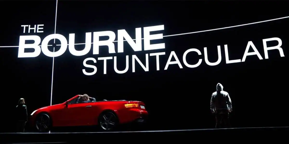 A red convertible car with two people beside it is on stage in front of a large, illuminated sign reading "The Bourne Stuntacular" in bold letters. The background is dark, making the sign stand out.