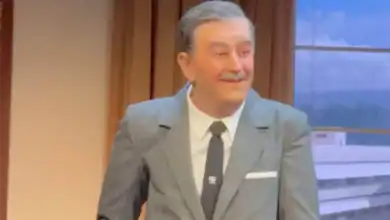 A wax figure of a smiling man with gray hair and a mustache, dressed in a gray suit, white shirt, and black tie, stands indoors near a window with a scenic backdrop outside.