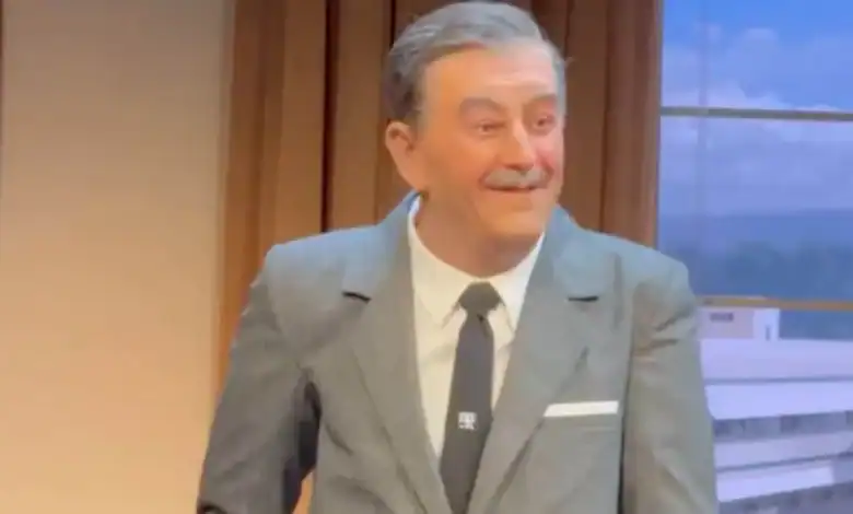 A wax figure of a smiling man with gray hair and a mustache, dressed in a gray suit, white shirt, and black tie, stands indoors near a window with a scenic backdrop outside.