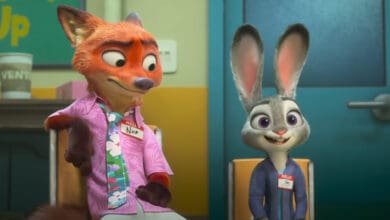 Nick and Judy, sit side by side in chairs at a meeting room. Both wear name tags and casual clothes, with Nick looking to the side and Judy smiling forward.