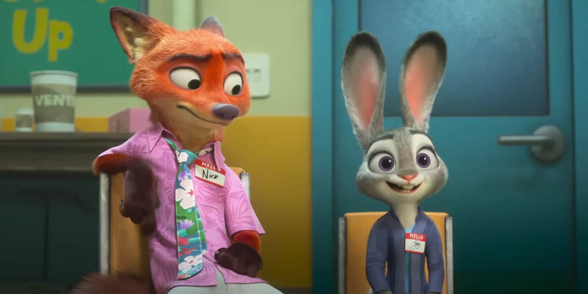 Nick and Judy, sit side by side in chairs at a meeting room. Both wear name tags and casual clothes, with Nick looking to the side and Judy smiling forward.
