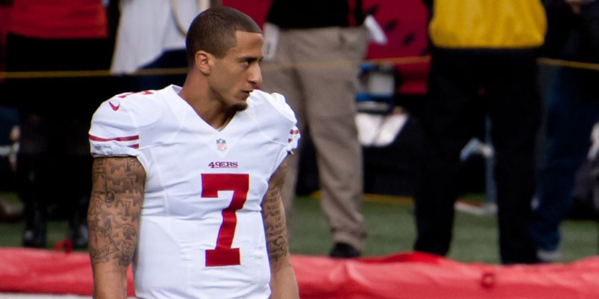 A football player wearing a white San Francisco 49ers uniform with the number 7 stands on the field, looking to the left. The background shows blurred people and equipment on the sidelines.