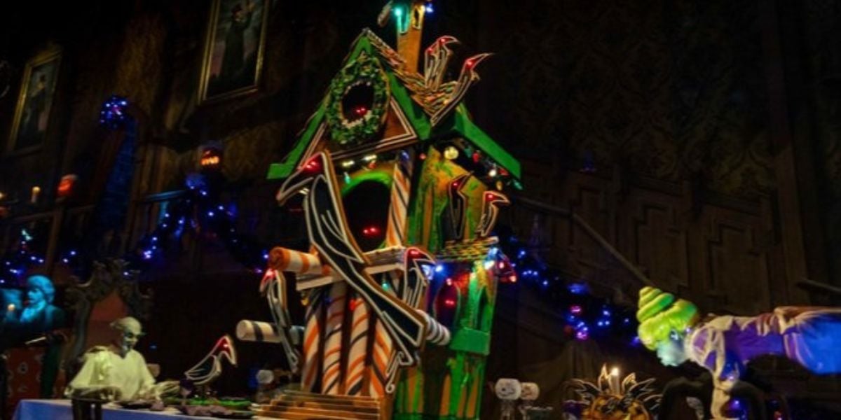 A spooky gingerbread house with monster arms stands on a table surrounded by Halloween decorations, glowing blue and green lights, and ghostly figures, creating a Haunted Mansion Holiday vibe in the dimly lit, haunted room.