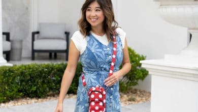 A woman smiles while walking outside, wearing a blue floral dress over a white shirt and carrying a small red and white polka dot crossbody bag. There are white columns, greenery, and outdoor chairs in the background.
