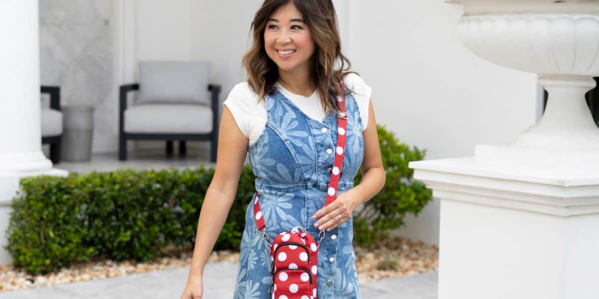 A woman smiles while walking outside, wearing a blue floral dress over a white shirt and carrying a small red and white polka dot crossbody bag. There are white columns, greenery, and outdoor chairs in the background.