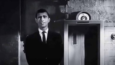 A man in a dark suit and tie stands in front of a door with a circular gauge above it, in a black-and-white setting with a textured wall in the background.