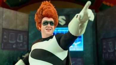 A person in a black and white superhero costume with a large red wig and black eye mask points forward and smiles, standing in front of futuristic screens and equipment.