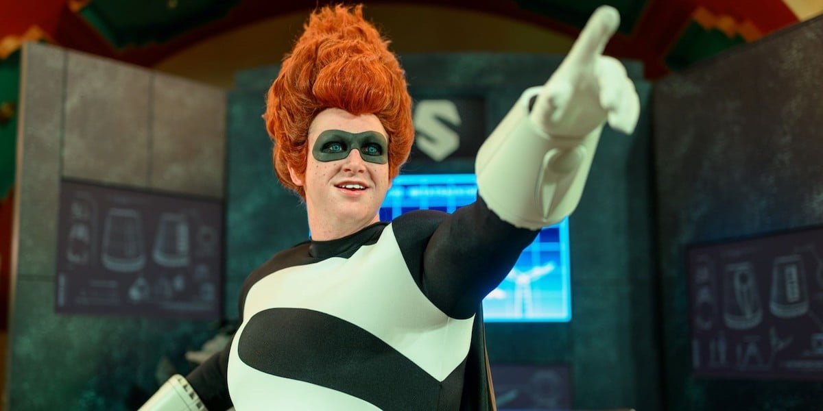 A person in a black and white superhero costume with a large red wig and black eye mask points forward and smiles, standing in front of futuristic screens and equipment.
