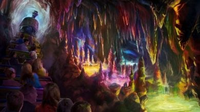 A group of people ride a train through a colorful, glowing cave filled with vibrant stalactites and stalagmites illuminated in shades of purple, orange, yellow, and green.
