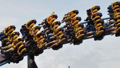People riding a roller coaster with bright yellow harnesses, legs dangling as the ride goes upside down on a loop against a cloudy sky. Riders have their arms raised and appear excited.