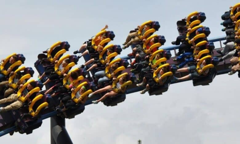 People riding a roller coaster with bright yellow harnesses, legs dangling as the ride goes upside down on a loop against a cloudy sky. Riders have their arms raised and appear excited.