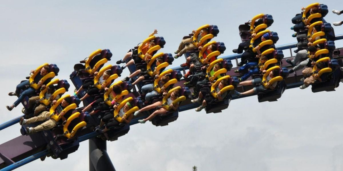 People riding a roller coaster with bright yellow harnesses, legs dangling as the ride goes upside down on a loop against a cloudy sky. Riders have their arms raised and appear excited.