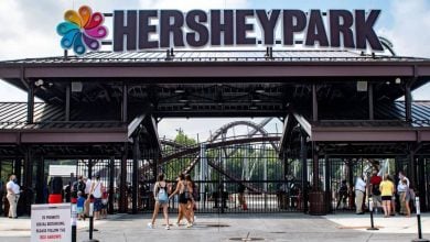 Entrance to Hersheypark with people walking in and out, large “HERSHEYPARK” sign overhead, a colorful logo, roller coaster tracks visible in the background, and a red information sign in front.