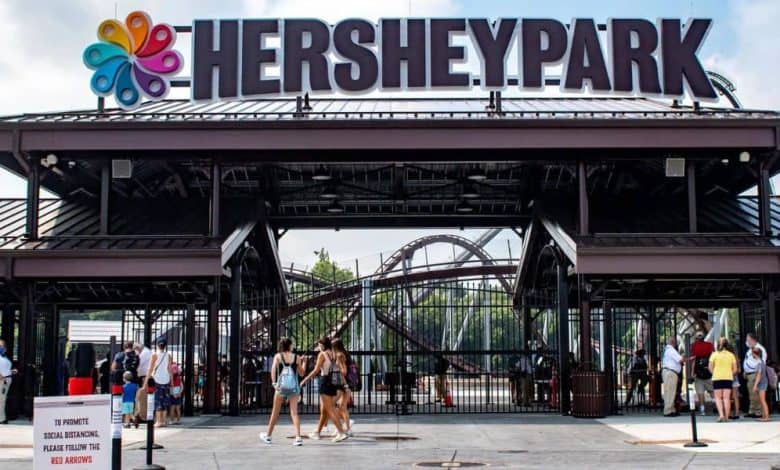 Entrance to Hersheypark with people walking in and out, large “HERSHEYPARK” sign overhead, a colorful logo, roller coaster tracks visible in the background, and a red information sign in front.