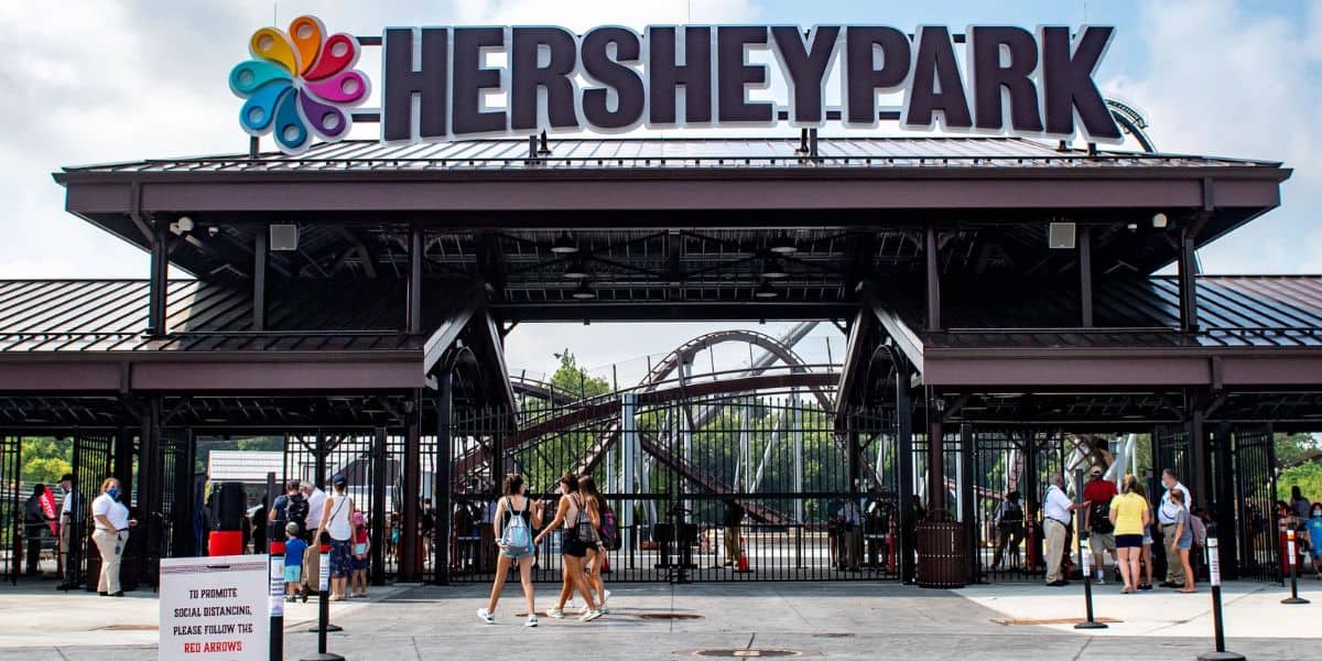 Entrance to Hersheypark with people walking in and out, large “HERSHEYPARK” sign overhead, a colorful logo, roller coaster tracks visible in the background, and a red information sign in front.