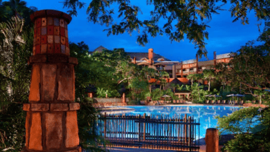 A large, circular outdoor swimming pool surrounded by lush trees and greenery at dusk, with a rustic stone pillar and wooden fence in the foreground and a warmly lit building in the background.