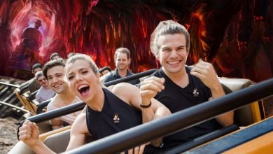A group of excited people ride a roller coaster through a dark, cave-like tunnel with glowing red and orange walls, smiling and raising their hands in joy.