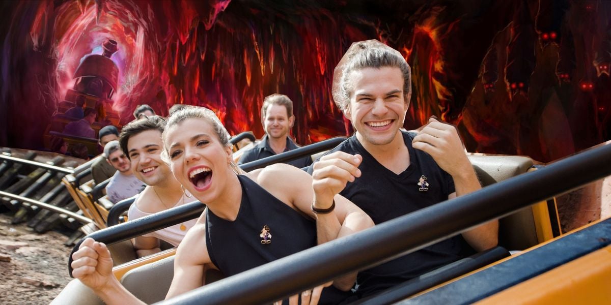A group of excited people ride a roller coaster through a dark, cave-like tunnel with glowing red and orange walls, smiling and raising their hands in joy.