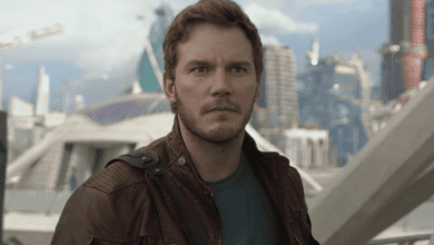 A man with short brown hair and a trimmed beard, wearing a maroon leather jacket and blue shirt, stands outdoors in a futuristic city with modern buildings in the background. He looks serious and focused.