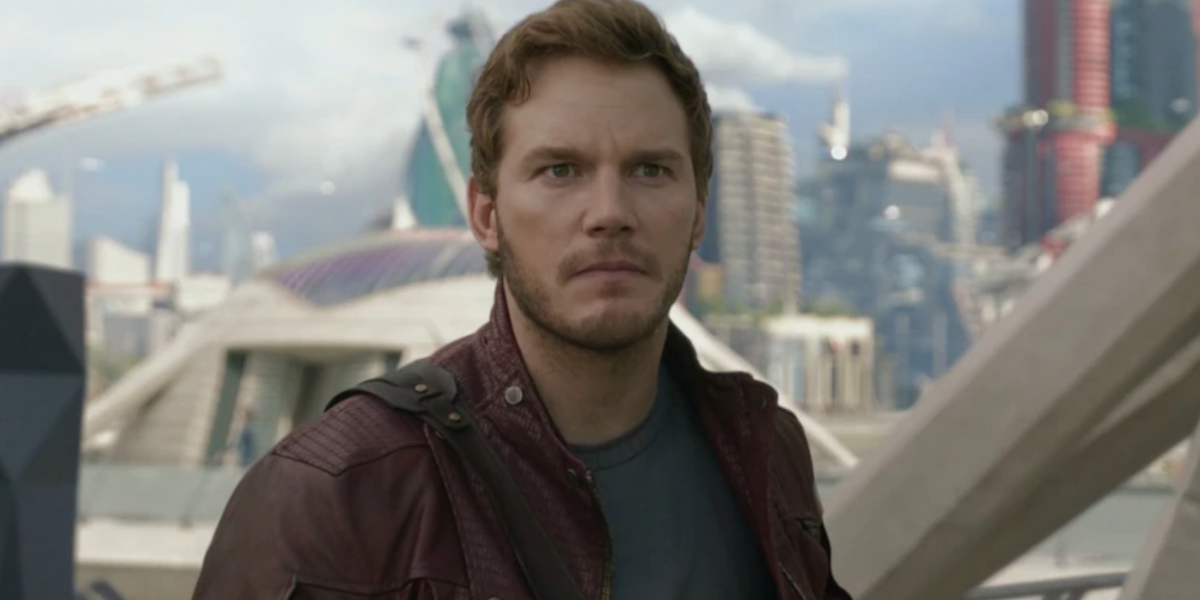 A man with short brown hair and a trimmed beard, wearing a maroon leather jacket and blue shirt, stands outdoors in a futuristic city with modern buildings in the background. He looks serious and focused.