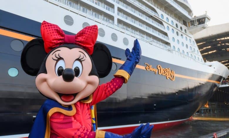 Minnie Mouse, dressed in a colorful outfit, poses and waves in front of a large cruise ship with the name "Disney Destiny" painted on its side. The ship is docked near a covered structure. Disney Cruise Line alcohol policy change