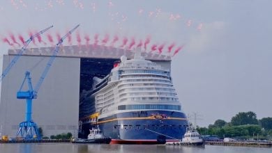 A large cruise ship is launched from a shipyard, guided by tugboats on a river, with red fireworks arching above. Blue cranes and a massive hangar are visible in the background.