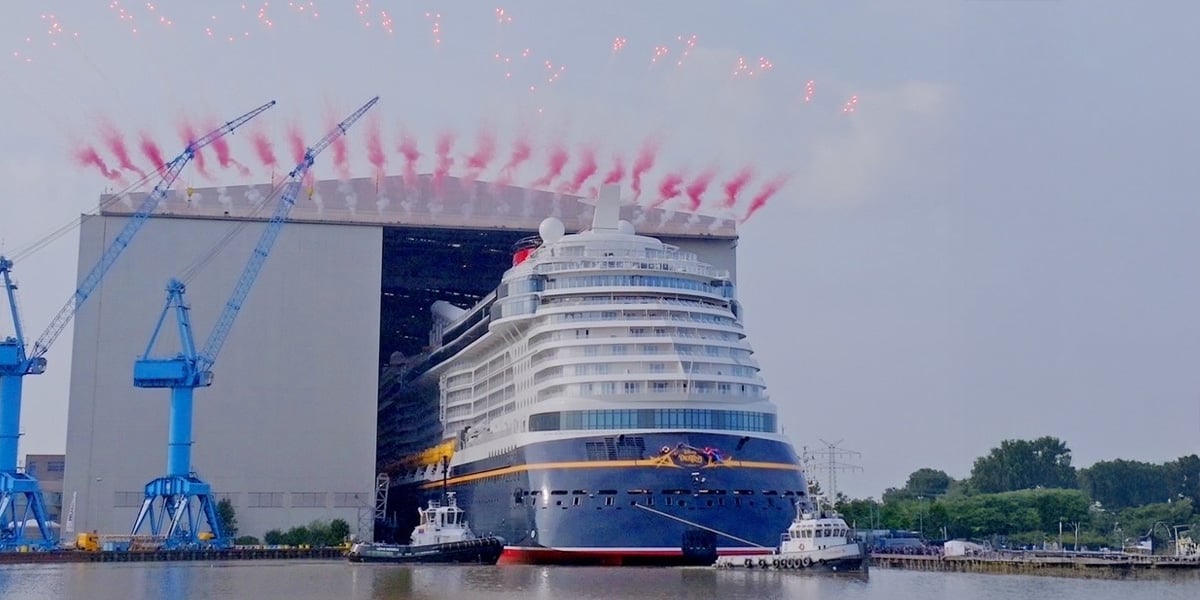 A large cruise ship is launched from a shipyard, guided by tugboats on a river, with red fireworks arching above. Blue cranes and a massive hangar are visible in the background.