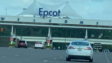 Cars approach the entrance gate to Epcot at Walt Disney World, with a large white canopy and "Epcot" sign above the word "WELCOME."