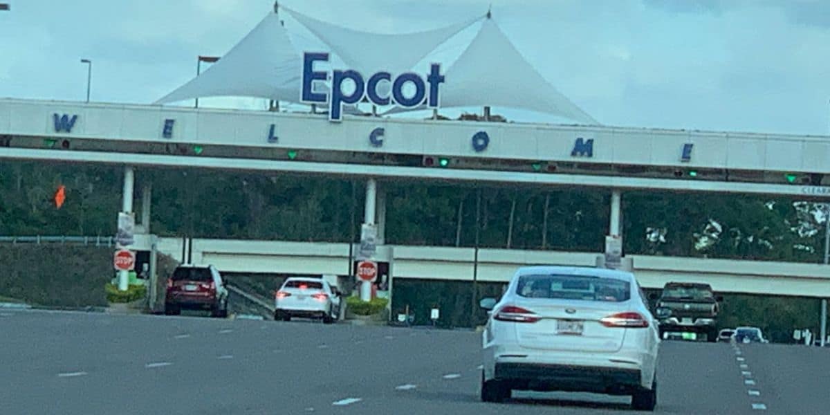 Cars approach the entrance gate to Epcot at Walt Disney World, with a large white canopy and "Epcot" sign above the word "WELCOME."